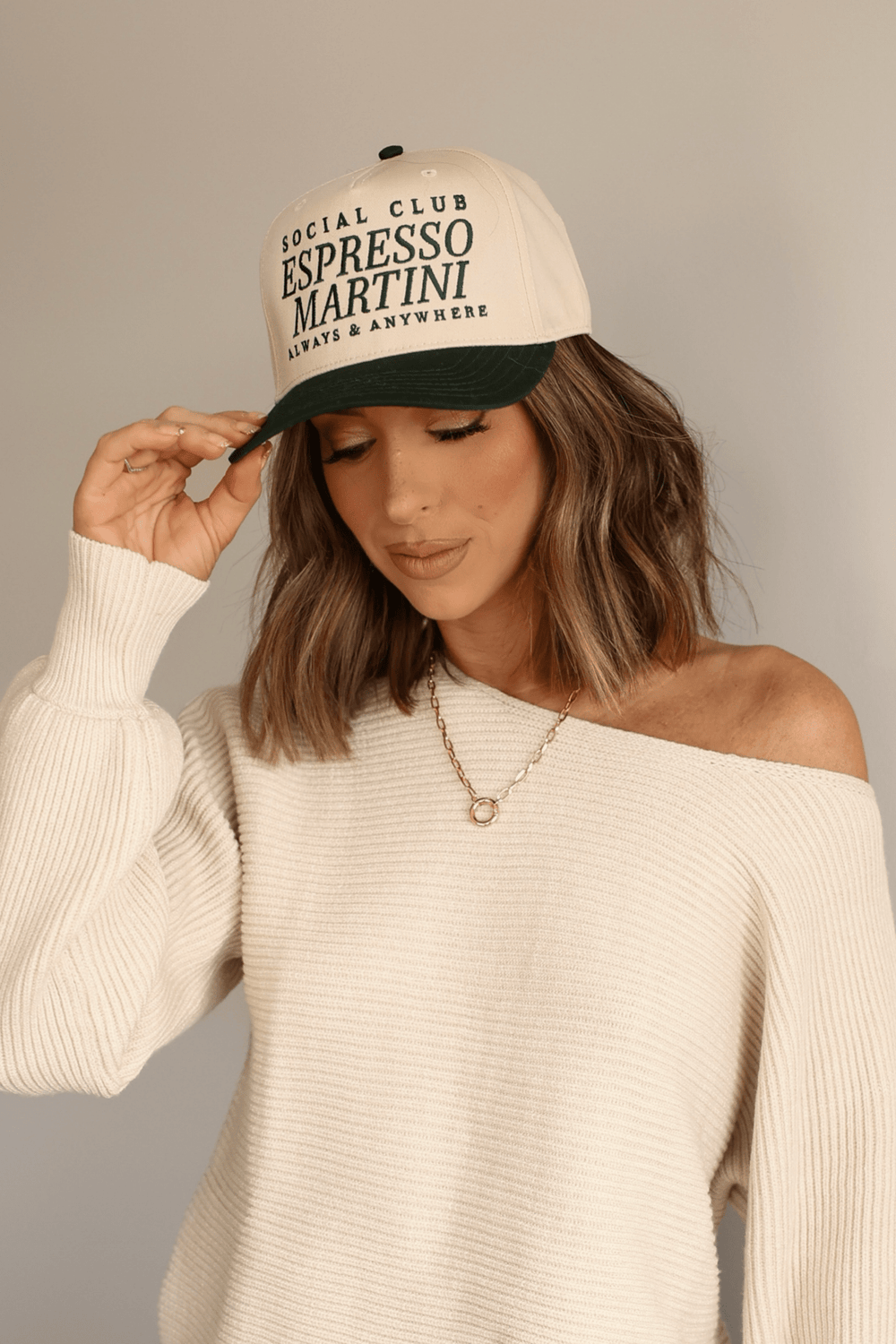 Woman in a cream sweater smiles, tilting her Green Social Club Espresso Martini Trucker Hat—perfect for coffee lovers.