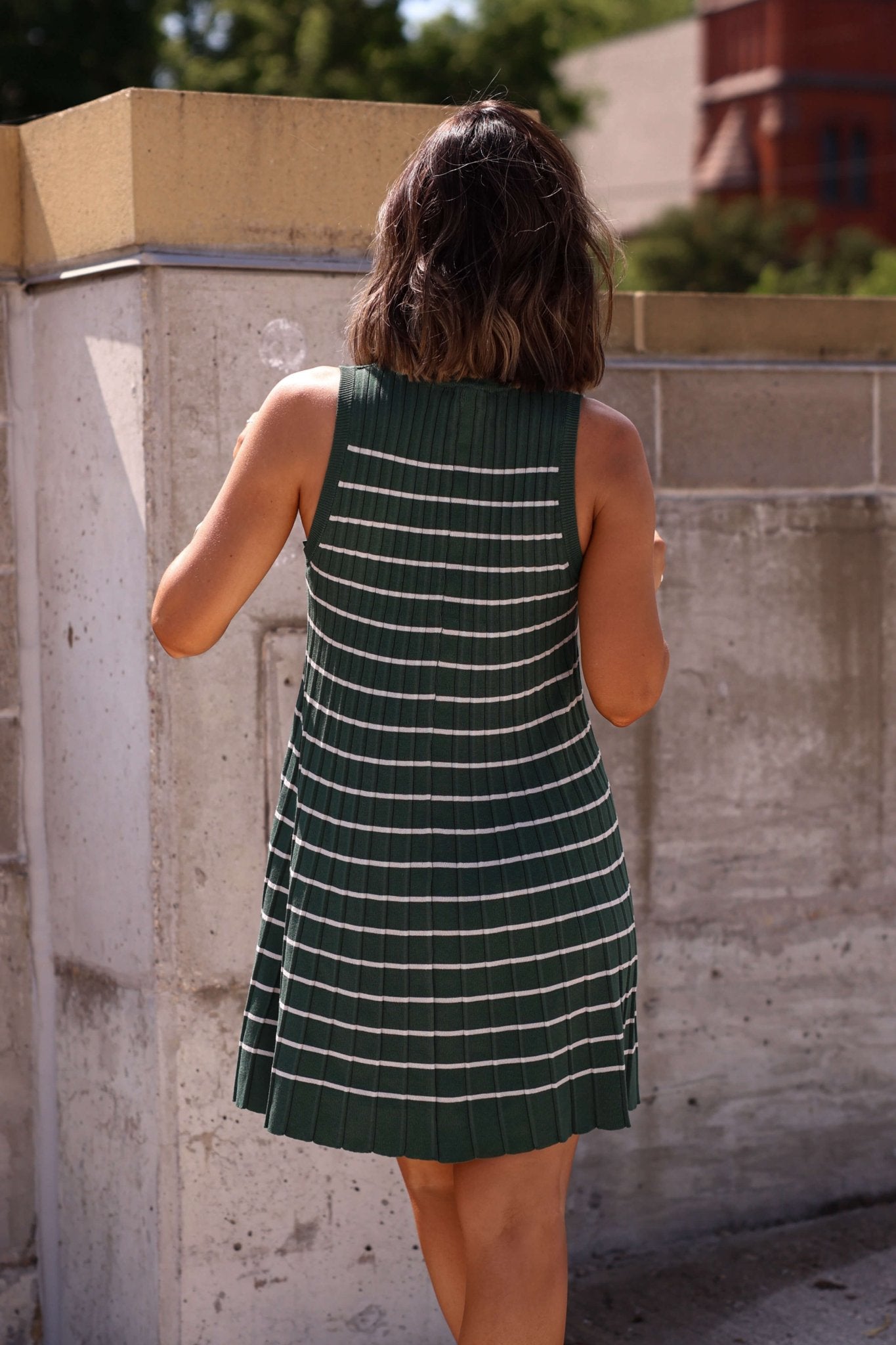 A woman in a Green Stripe Pleated Mini Dress - FINAL SALE stands outside, her shoulder-length hair framing her by a concrete wall.