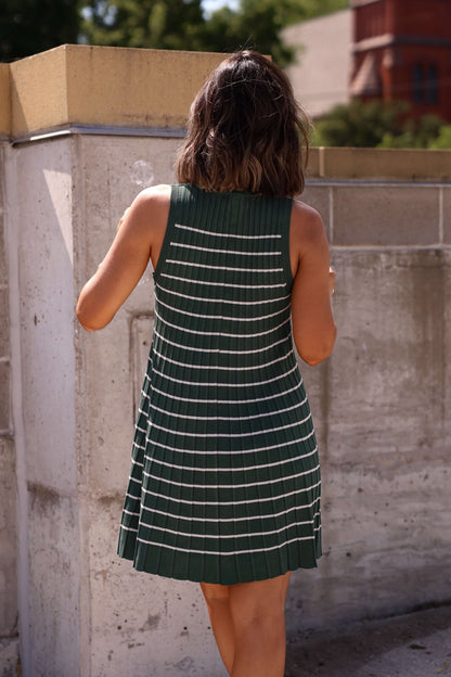 A woman in a Green Stripe Pleated Mini Dress - FINAL SALE stands outside, her shoulder-length hair framing her by a concrete wall.