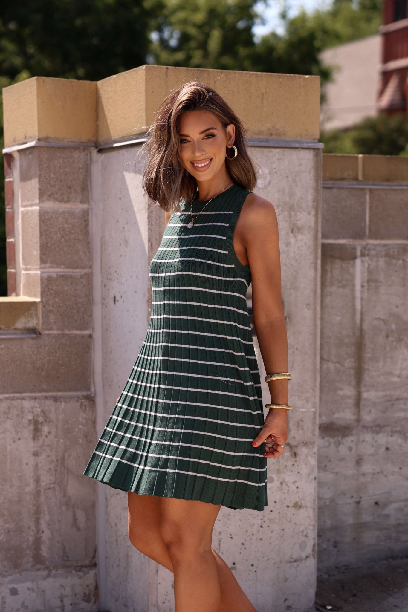 Woman with shoulder-length brown hair smiles in a Green Stripe Pleated Mini Dress, standing outside by a concrete wall.