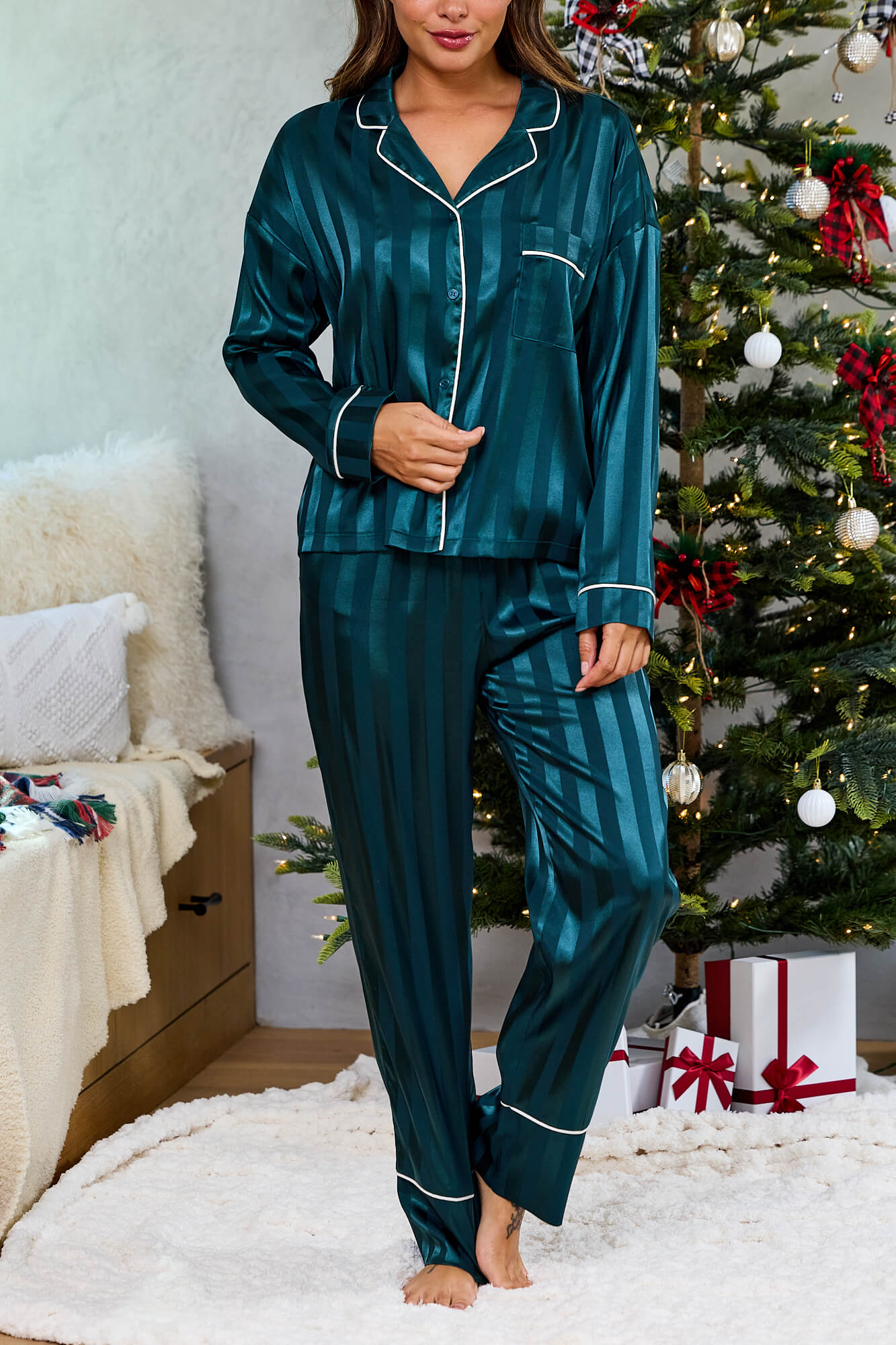 Barefoot woman in Green Striped Satin Pajama Set stands by a decorated Christmas tree with gifts and a cozy bed nearby.