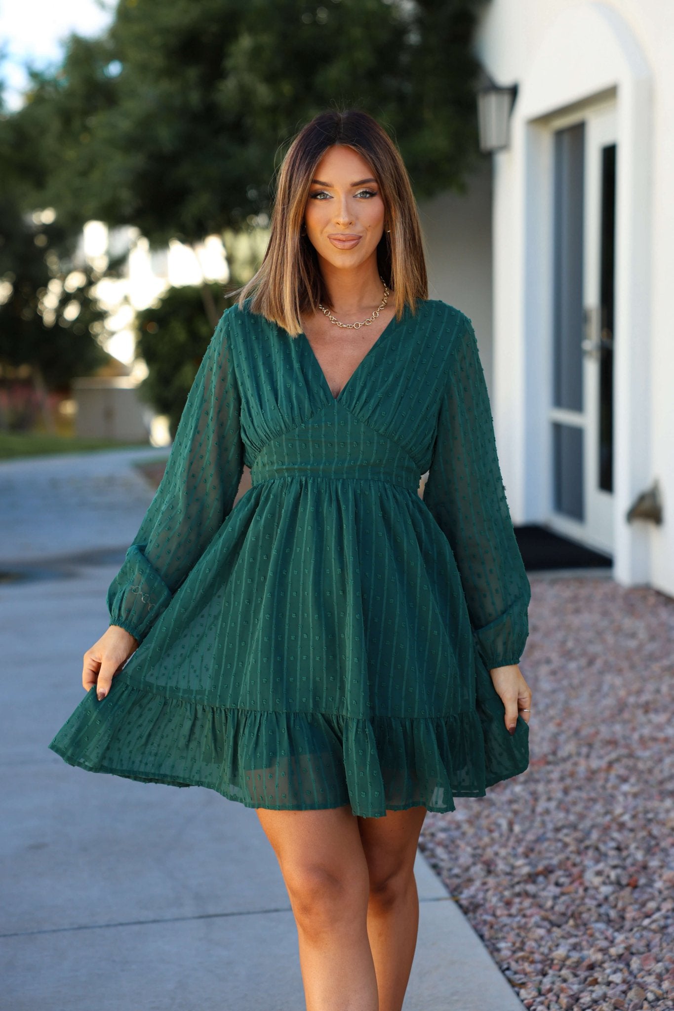 A woman models the Green Swiss Dot Ruffled Mini Dress outdoors, smiling and holding out the skirt for the camera.