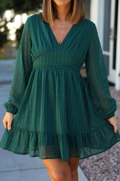 Woman models a Green Swiss Dot Ruffled Mini Dress with sheer long sleeves and ruffled hem, posing outdoors.