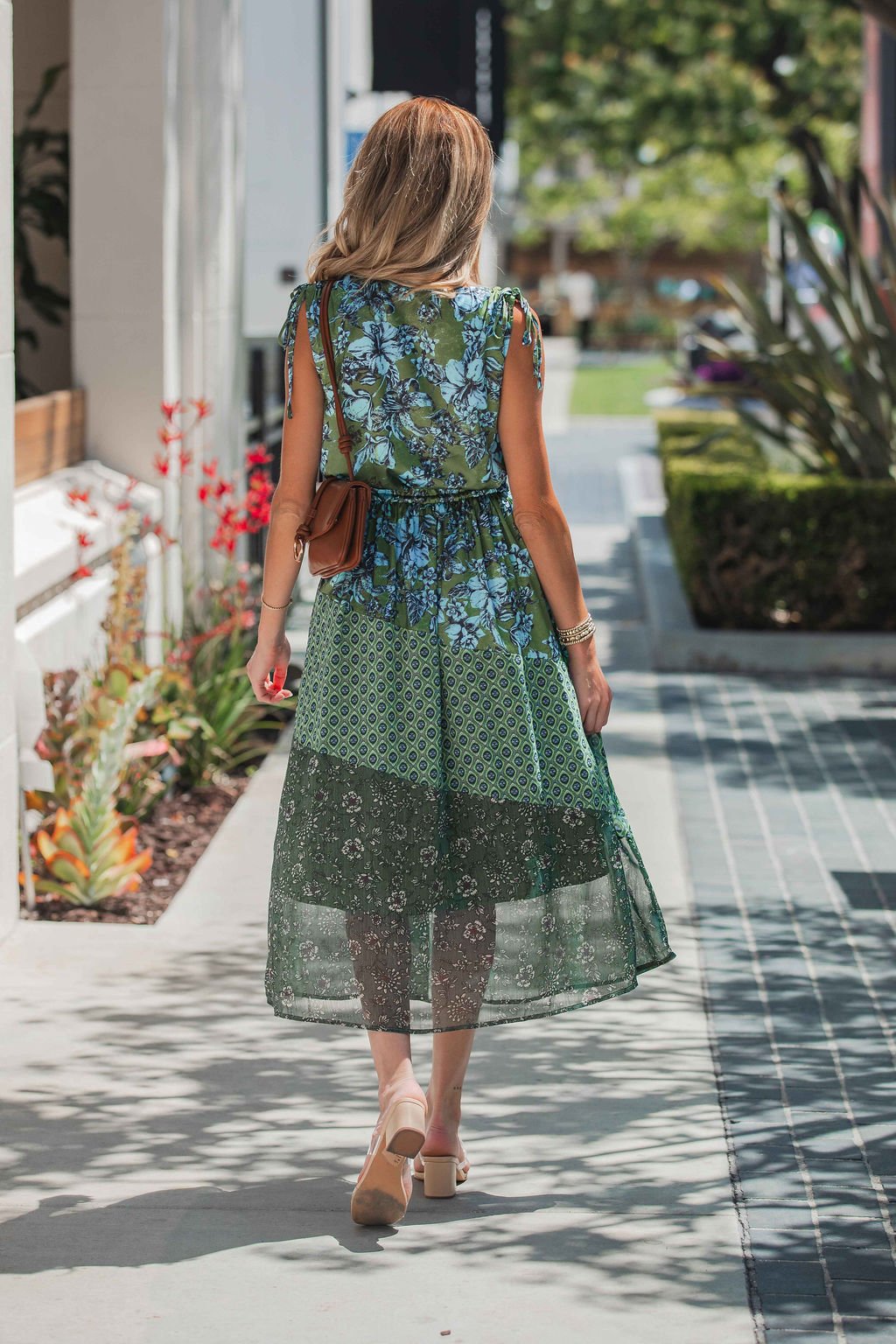 Wearing a Greenhaven Print Midi Dress, a woman walks with a brown purse and sandals along a sunny sidewalk with plants nearby.