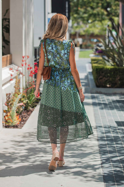 Wearing a Greenhaven Print Midi Dress, a woman walks with a brown purse and sandals along a sunny sidewalk with plants nearby.
