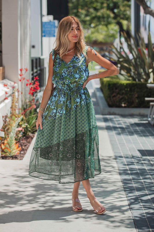 Woman in a Greenhaven Print Midi Dress stands on a sunny sidewalk, surrounded by greenery, looking to the side.