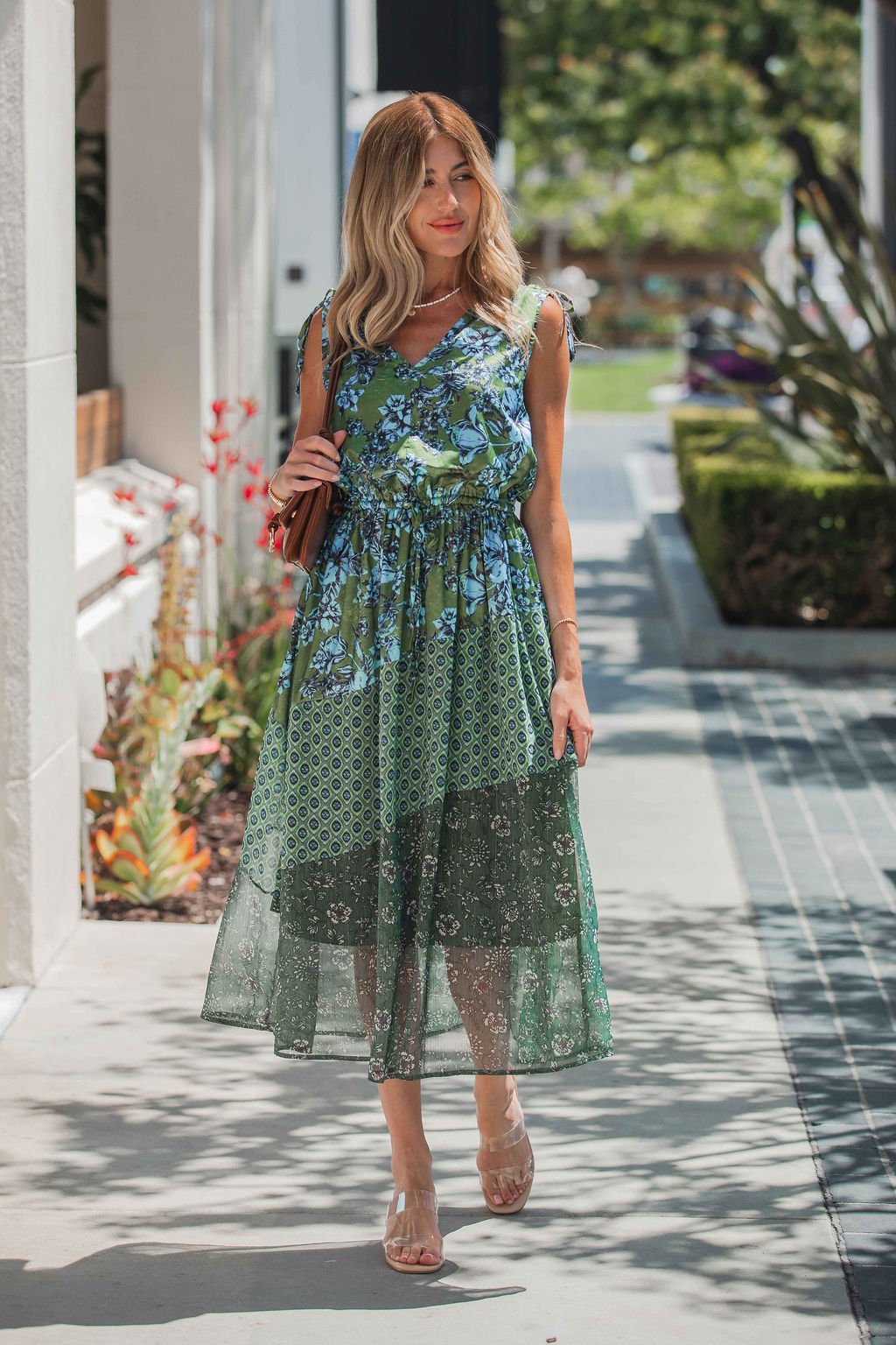 A woman in a Greenhaven Print Midi Dress walks outside on a sunny day with a brown handbag and clear sandals.
