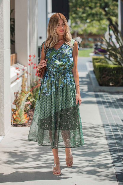 A woman in a Greenhaven Print Midi Dress walks outside on a sunny day with a brown handbag and clear sandals.