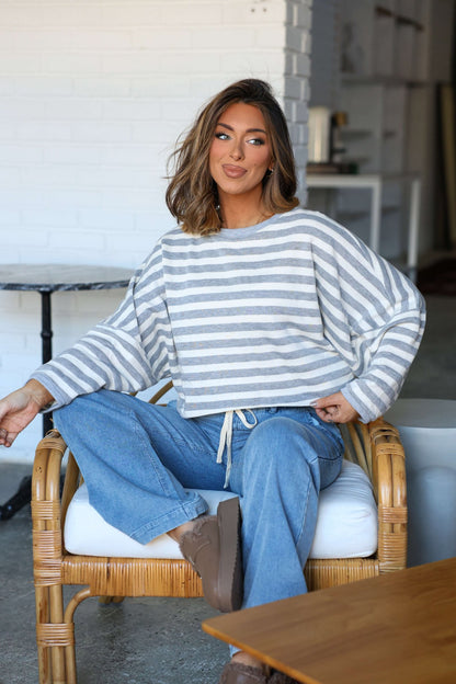 A woman sits indoors on a wicker chair, wearing a Grey and White Striped Boxy Pullover and blue jeans, looking relaxed.