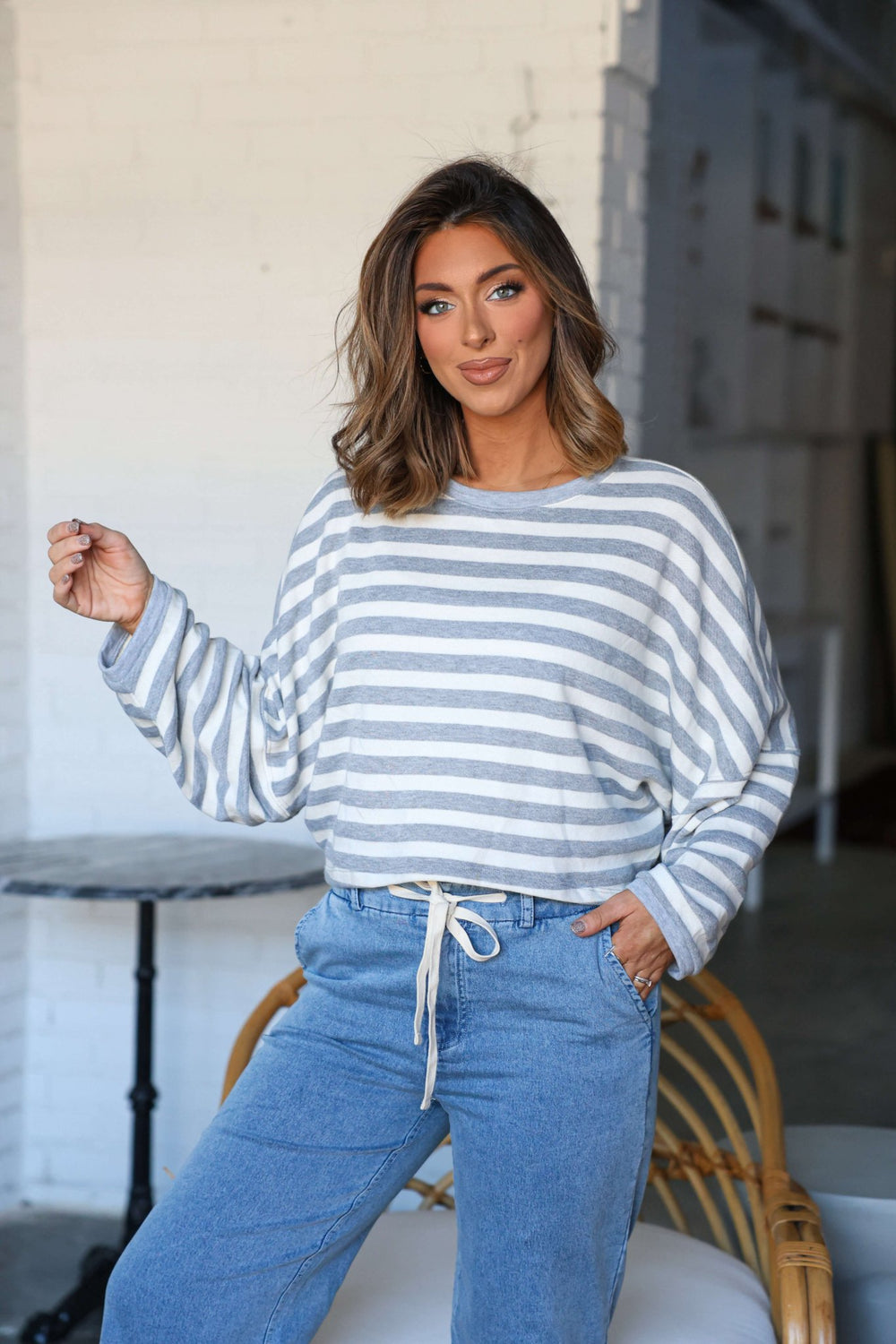Woman with wavy hair smiles at the camera indoors, wearing a Grey and White Striped Boxy Pullover and blue jeans.