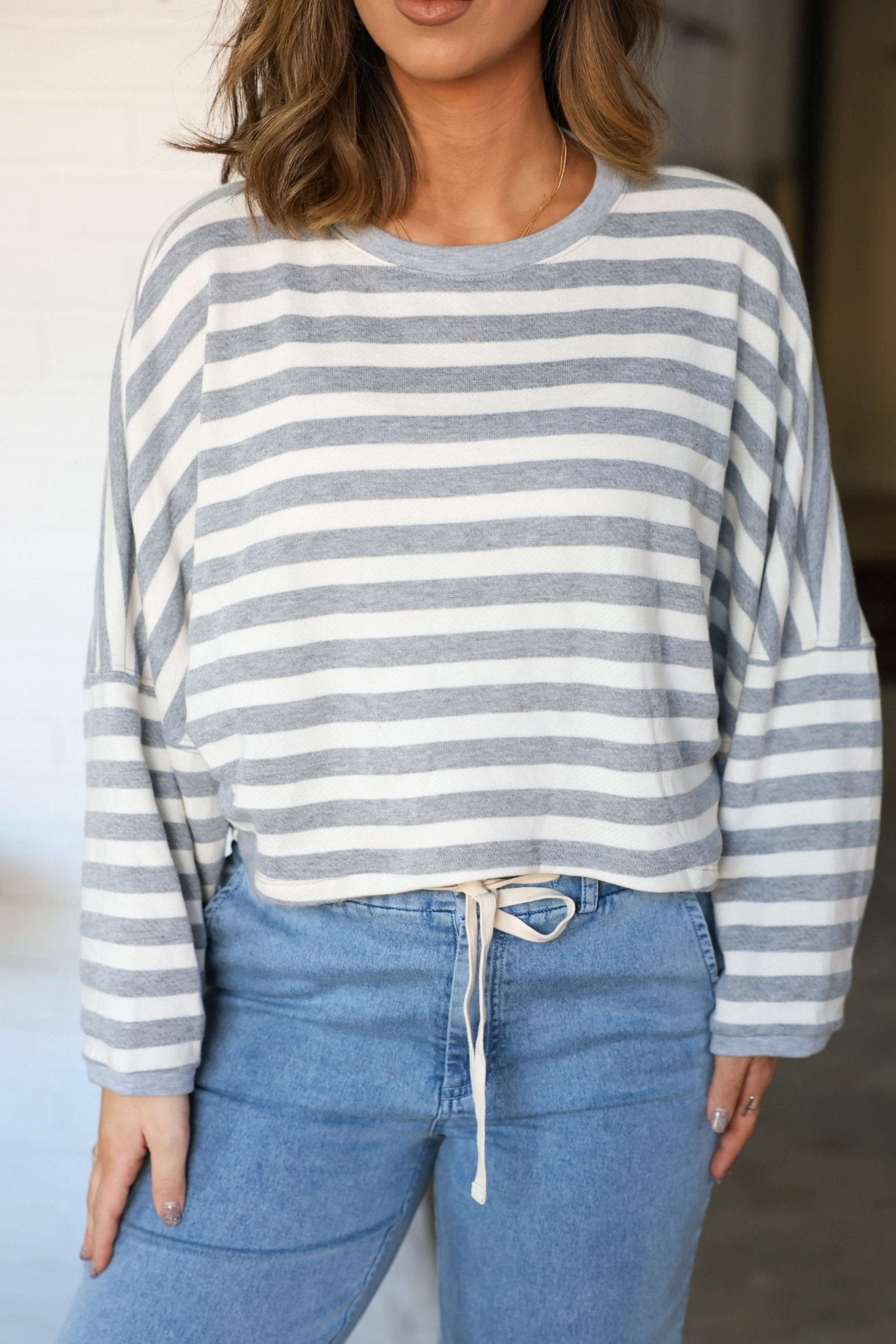 Woman wears a Grey and White Striped Boxy Pullover tucked into high-waisted blue jeans for a relaxed, drawstring-waist silhouette.