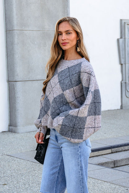Woman in a grey argyle print sweater and light jeans stands outside by a gray building, holding a black purse and smiling slightly.