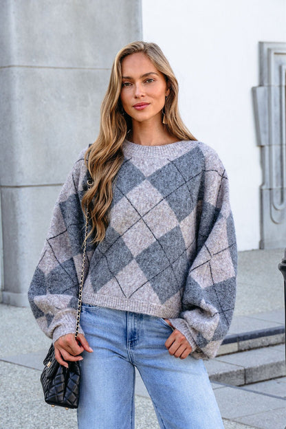 Woman with long hair wears a Grey Argyle Print Sweater, light blue jeans, and holds a quilted black purse outdoors.