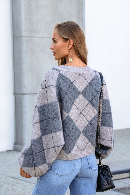 Woman with long hair wears a Grey Argyle Print Sweater, blue jeans, statement earrings, and carries a black quilted purse outdoors.