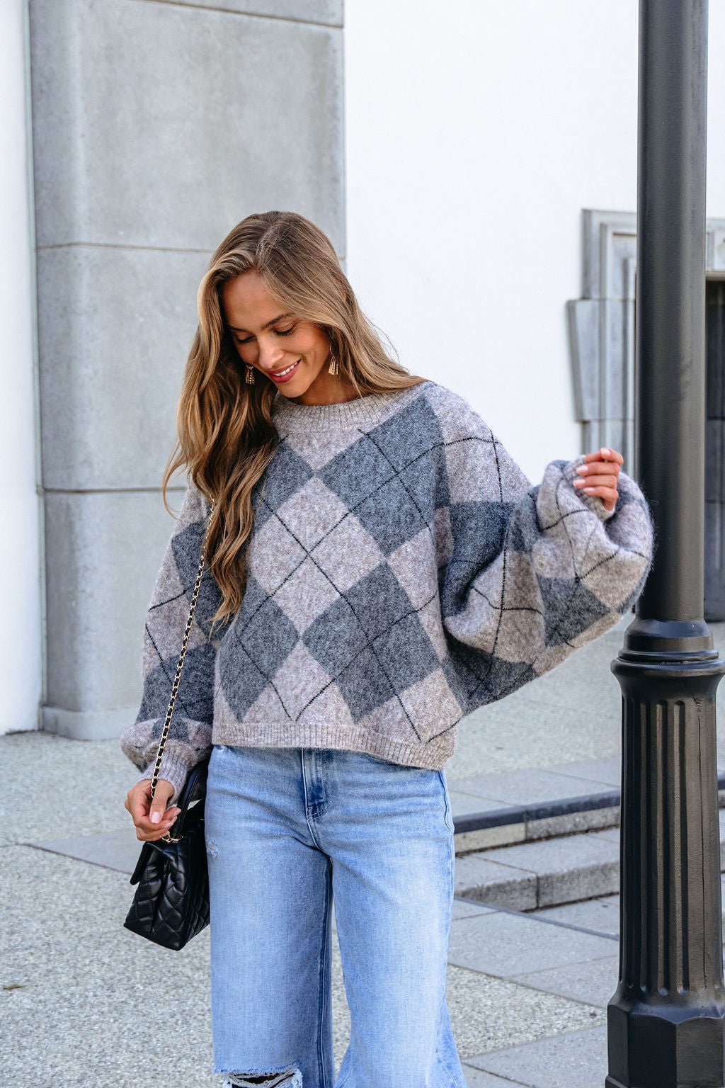 Woman wears a Grey Argyle Print Sweater and blue jeans, smiling by a black lamp post while holding a black purse.