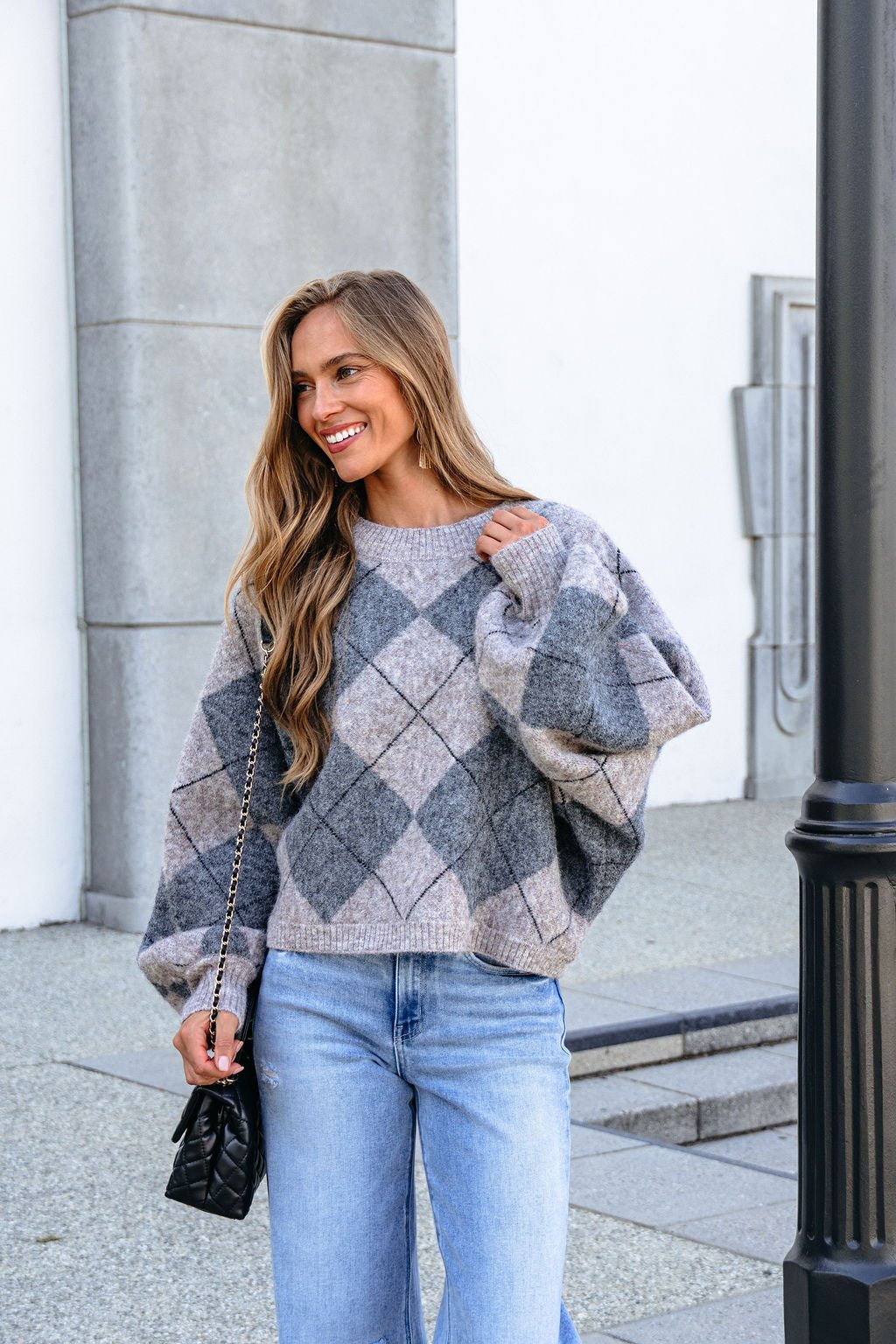 Woman in a Grey Argyle Print Sweater and jeans smiles outdoors, holding a black quilted purse.