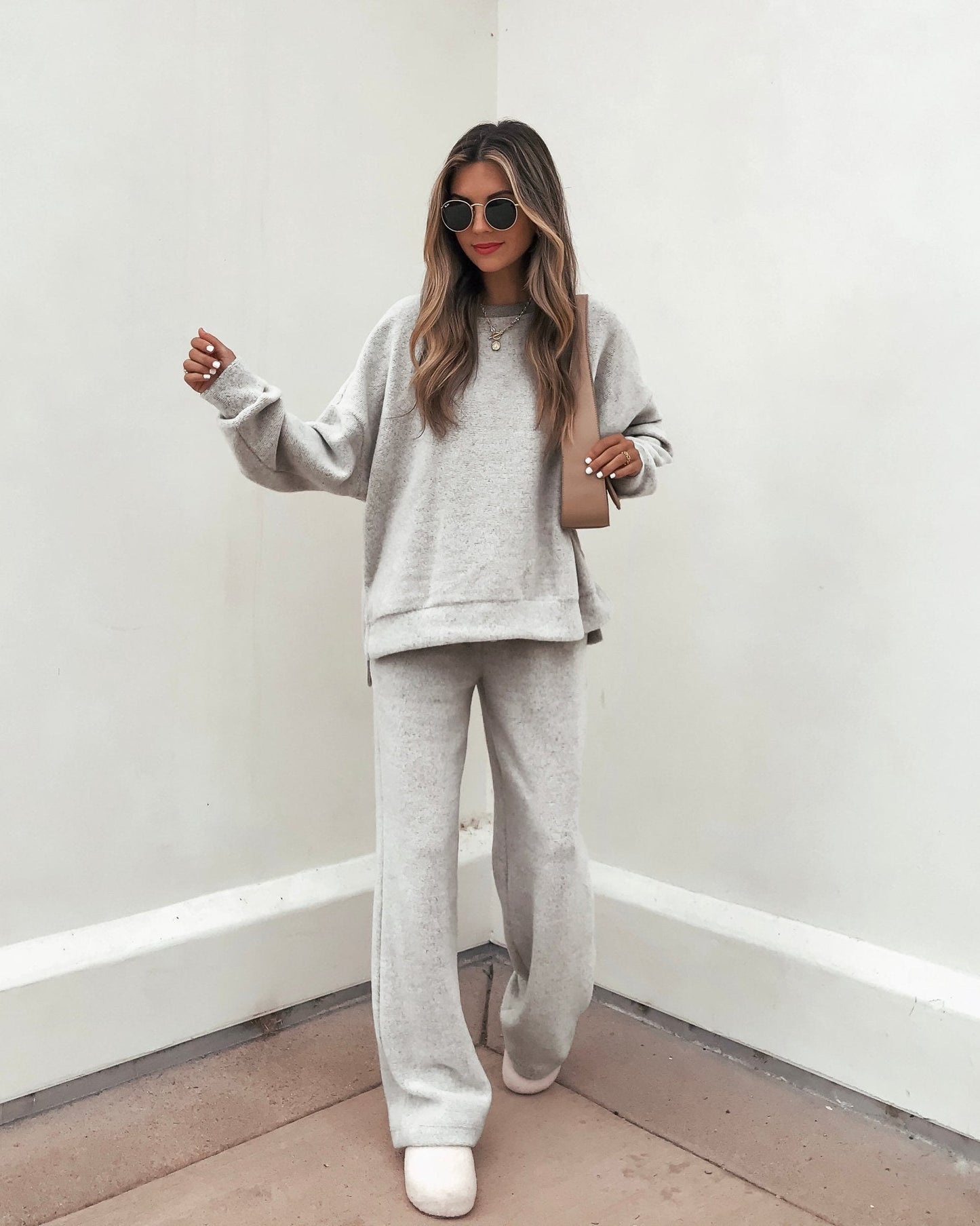 Woman in an oversized light gray sweater and Grey Brushed Lounge Sweatpants, holding a small purse by a white wall.