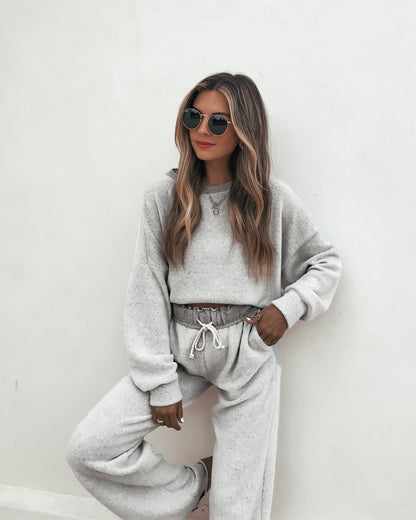 Woman in Grey Brushed Lounge Sweatpants with sunglasses poses by a white wall, hand on hip, her hair loose and wavy.