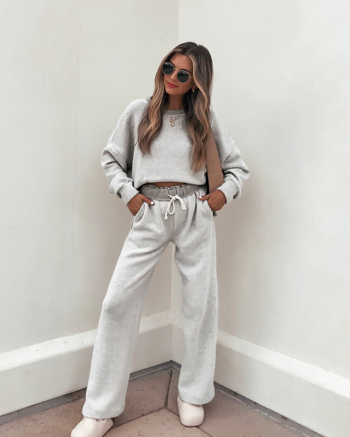 Woman in a Grey Brushed Lounge Sweatshirt with a relaxed fit, sunglasses, and slippers, standing against a white wall.