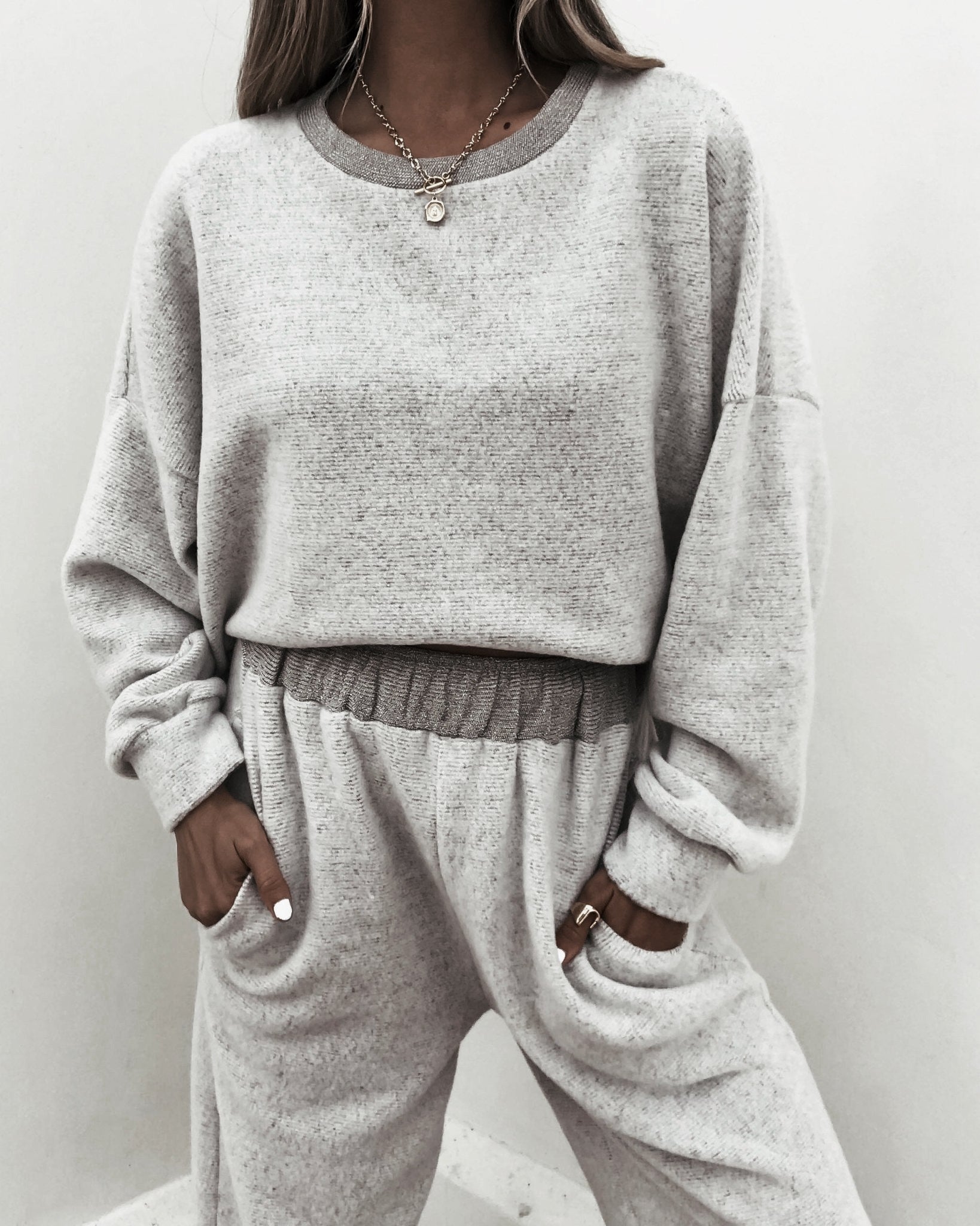 A woman wears a Grey Brushed Lounge Sweatshirt and sweatpants, styled with layered necklaces and rings.