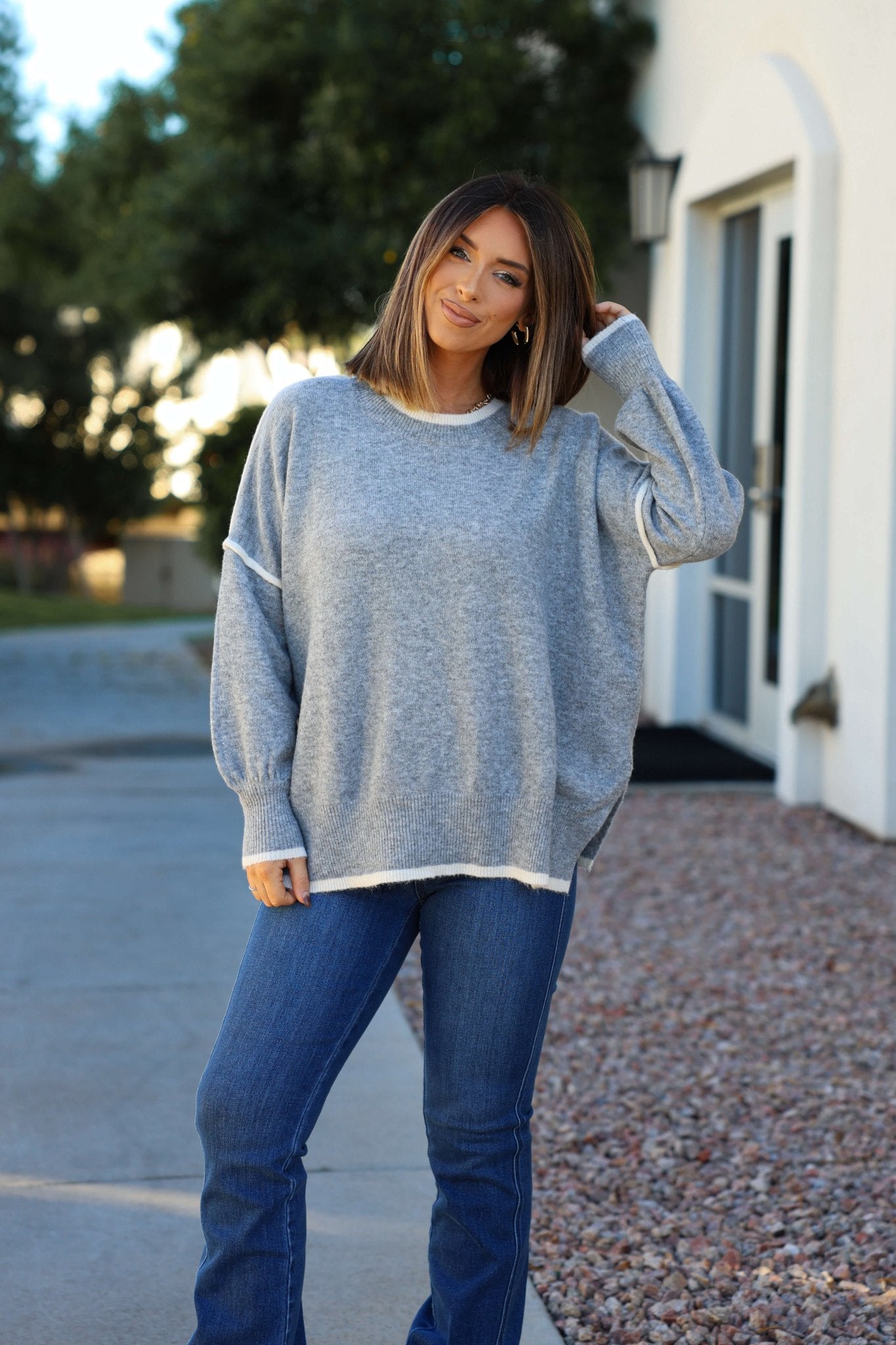 A woman in a Grey Contrast Detail Pullover Sweater and blue jeans smiles outside—an ideal look for fall.