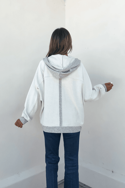 A person wears a Grey Contrast Half Button Up Hooded Sweatshirt and blue jeans, standing facing a white wall with arms slightly bent.