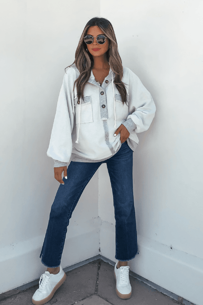 Woman in sunglasses wears a Grey Contrast Half Button Up Hooded Sweatshirt, blue jeans, and white sneakers against a white wall.