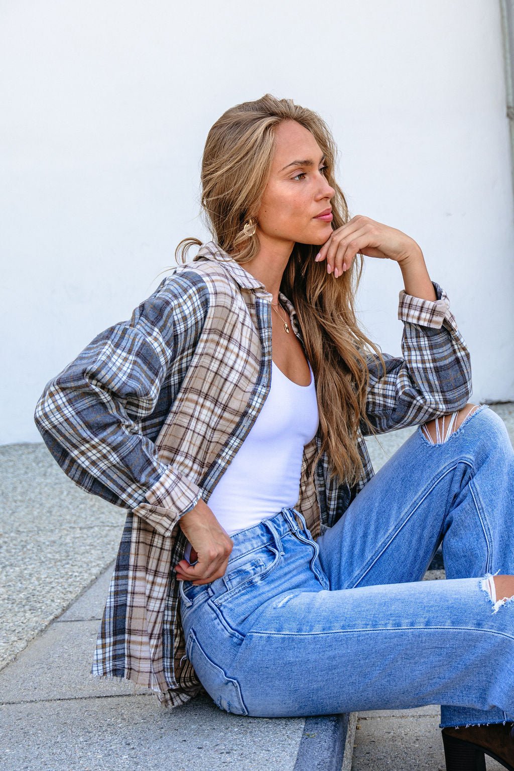 Woman with long hair sits on a step in a Grey Plaid Button Down Top, white tank, and ripped jeans, gazing thoughtfully aside.
