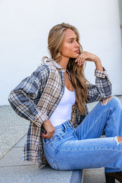 Woman with long hair sits on a step in a Grey Plaid Button Down Top, white tank, and ripped jeans, gazing thoughtfully aside.