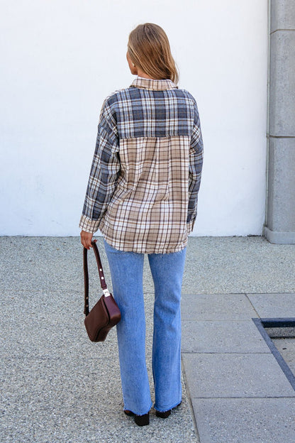 A person in a Grey Plaid Button Down Top, blue jeans, and holding a brown bag stands before a stone wall.