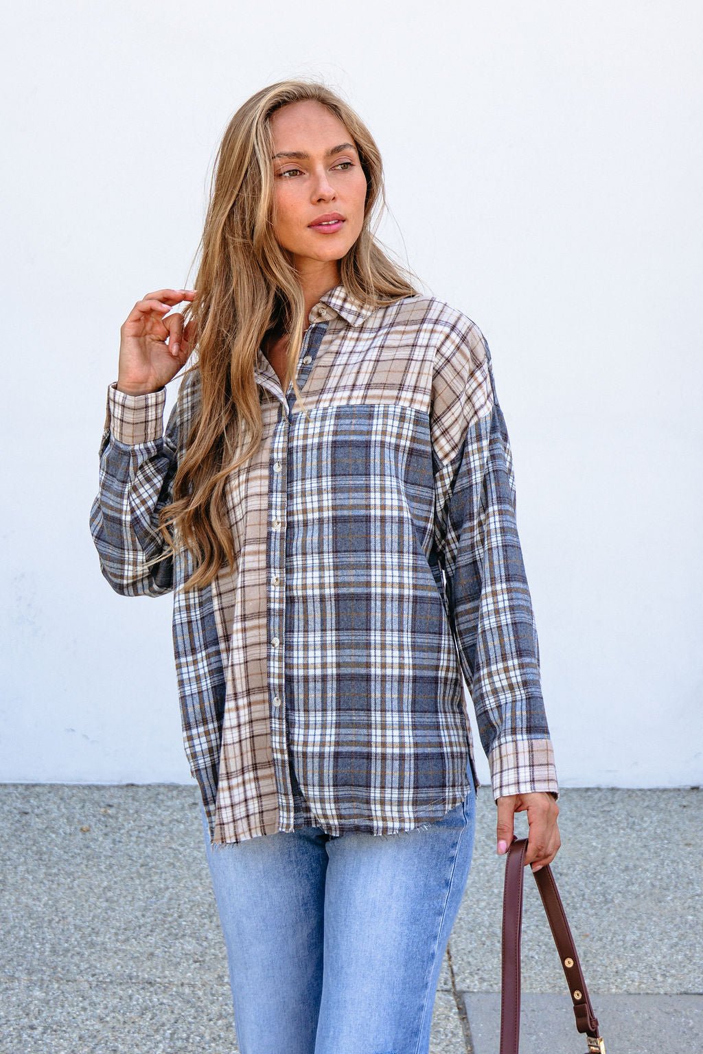Woman wears a Grey Plaid Button Down Top and light blue jeans outside, holding a brown strap with a white wall behind her.