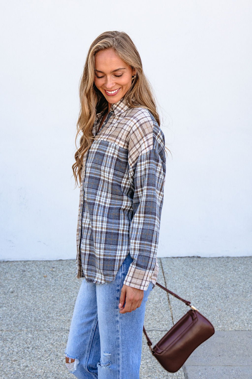Woman in relaxed fit Grey Plaid Button Down Top and ripped jeans smiles outside, holding a brown handbag against a white wall.