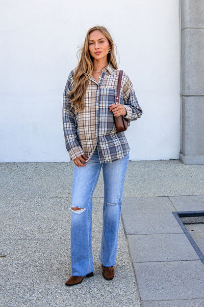 Woman in a Grey Plaid Button Down Top, ripped jeans, and brown shoes stands on a sidewalk with a simple background.