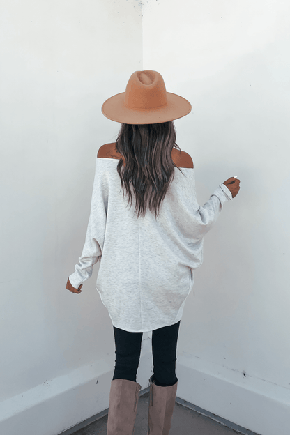 Woman with long wavy hair wears a Grey Soft Cloud Scuba Pullover, tan hat, black pants, and boots against a white wall.
