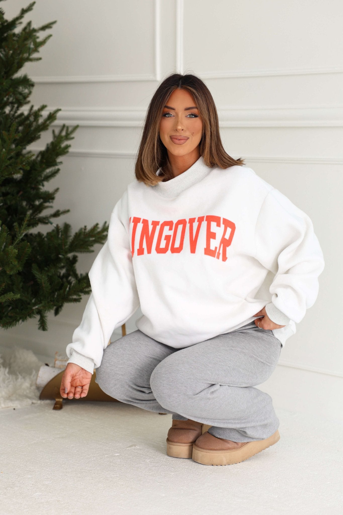 Woman squatting by a Christmas tree in a cozy Grey Spicy Margs Reversible Sweatshirt, gray pants, and tan slippers.