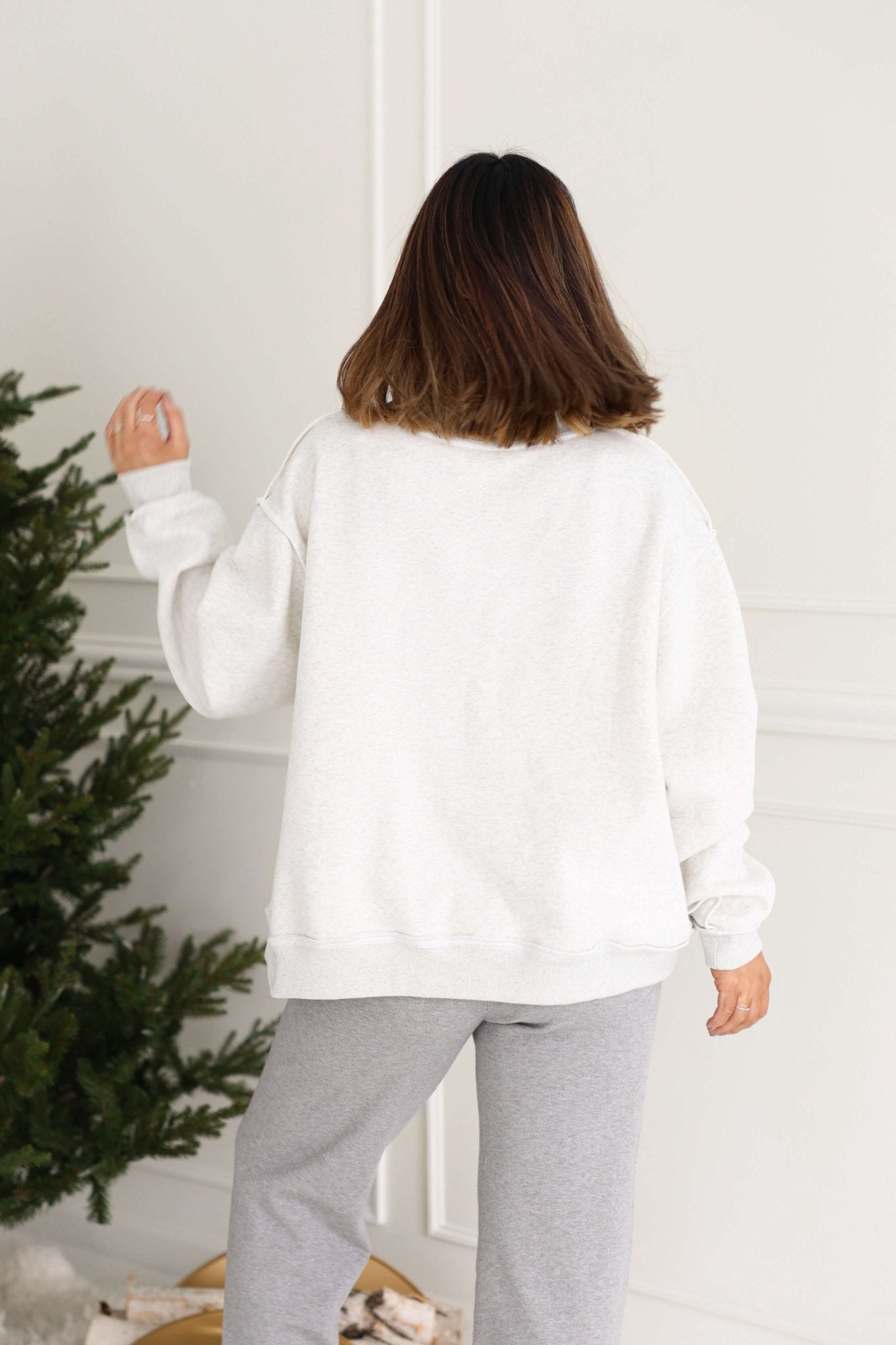Wearing the Grey Spicy Margs Reversible Sweatshirt, a person stands by a Christmas tree with their back to the camera.