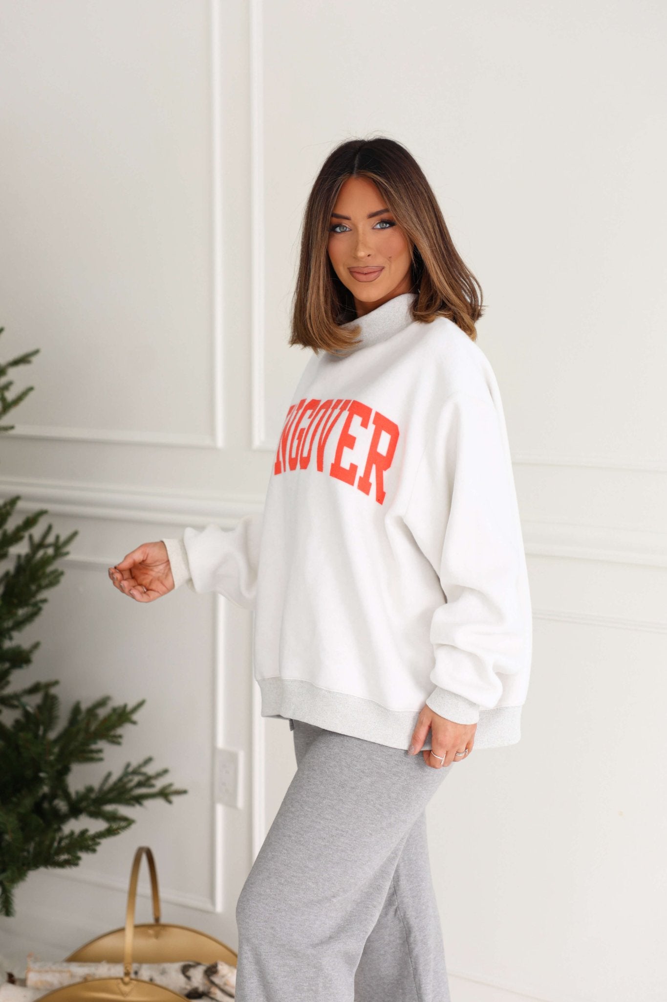 A woman with shoulder-length brown hair wears the Grey Spicy Margs Reversible Sweatshirt and gray pants by a green plant indoors.