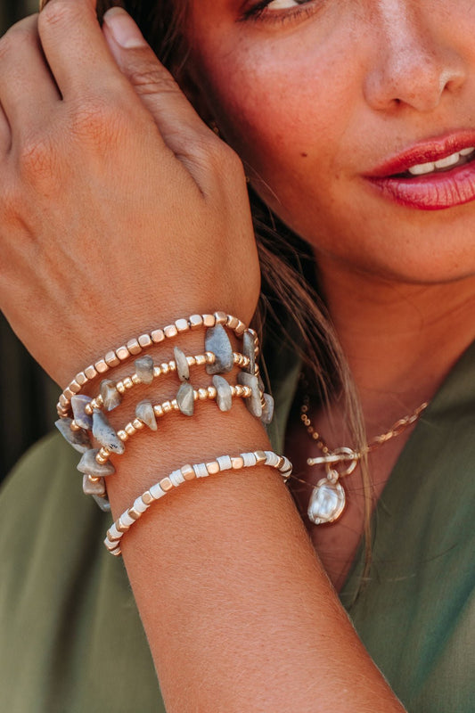 A woman in a green top touches her face, wearing the Grey Stone Beaded Bracelet Set - DOORBUSTER and a pearl necklace.
