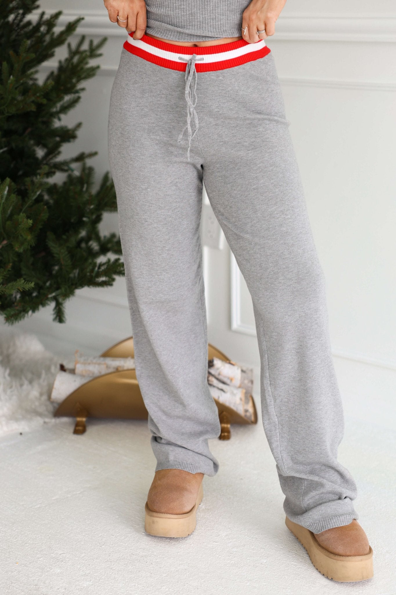 Person in Grey Striped Drawstring Pants and tan boots stands by a decorated tree and logs on a white carpet.