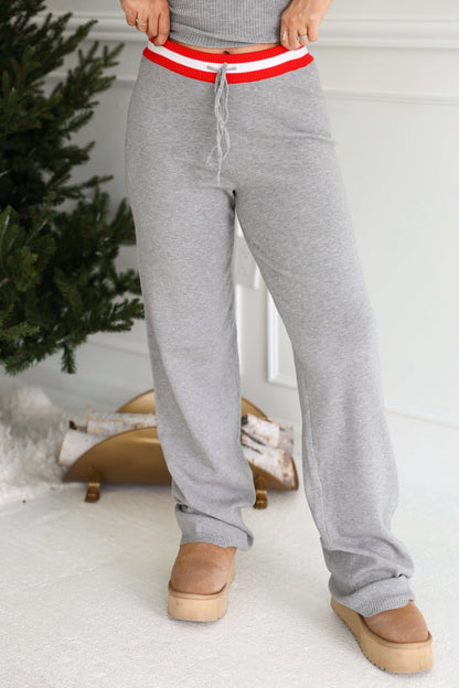 Person in Grey Striped Drawstring Pants and tan boots stands by a decorated tree and logs on a white carpet.