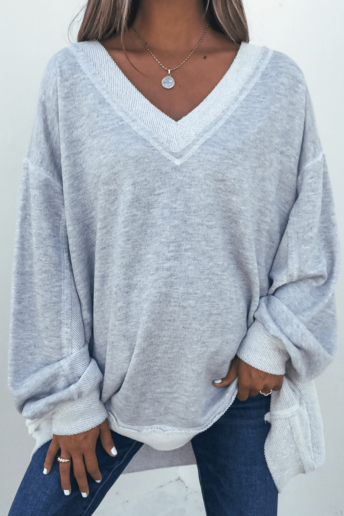 A person wears a Grey Two Tone Oversized Sweatshirt, blue jeans, and a necklace, with one hand in their pocket.