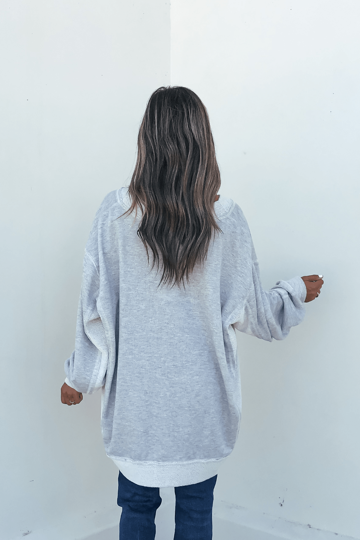A person with long wavy hair in a Grey Two Tone Oversized Sweatshirt and jeans stands facing a blank white wall.