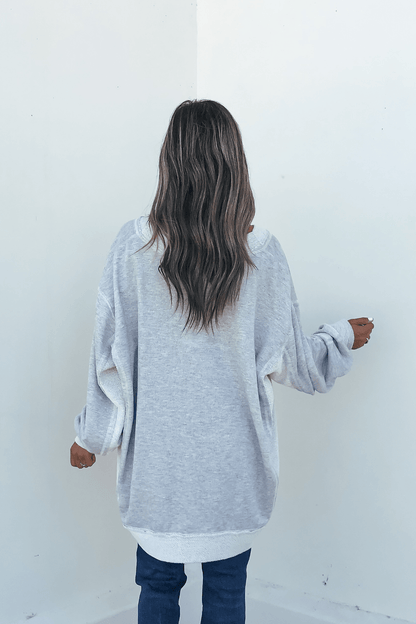 A person with long wavy hair in a Grey Two Tone Oversized Sweatshirt and jeans stands facing a blank white wall.