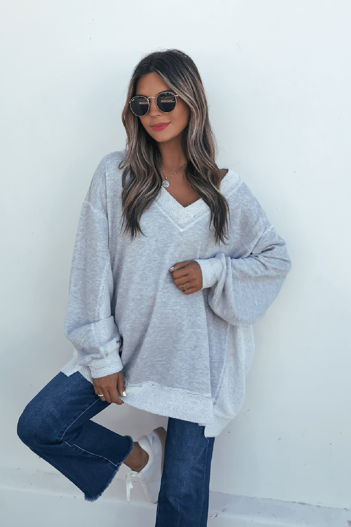 Woman wearing sunglasses, a Grey Two Tone Oversized Sweatshirt, blue jeans, and white sneakers poses casually against a white wall.