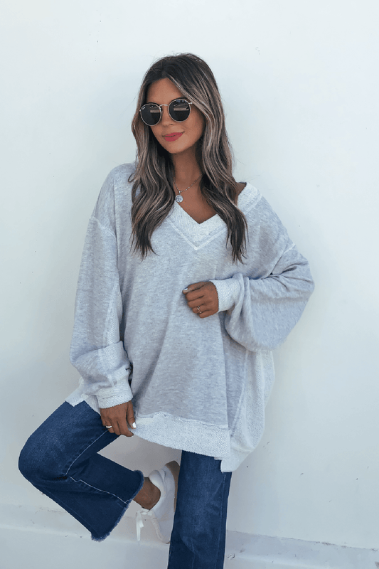 Woman wearing sunglasses, a Grey Two Tone Oversized Sweatshirt, blue jeans, and white sneakers poses casually against a white wall.