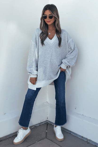 Woman in sunglasses, Grey Two Tone Oversized Sweatshirt, blue jeans, and white sneakers stands against a white wall.