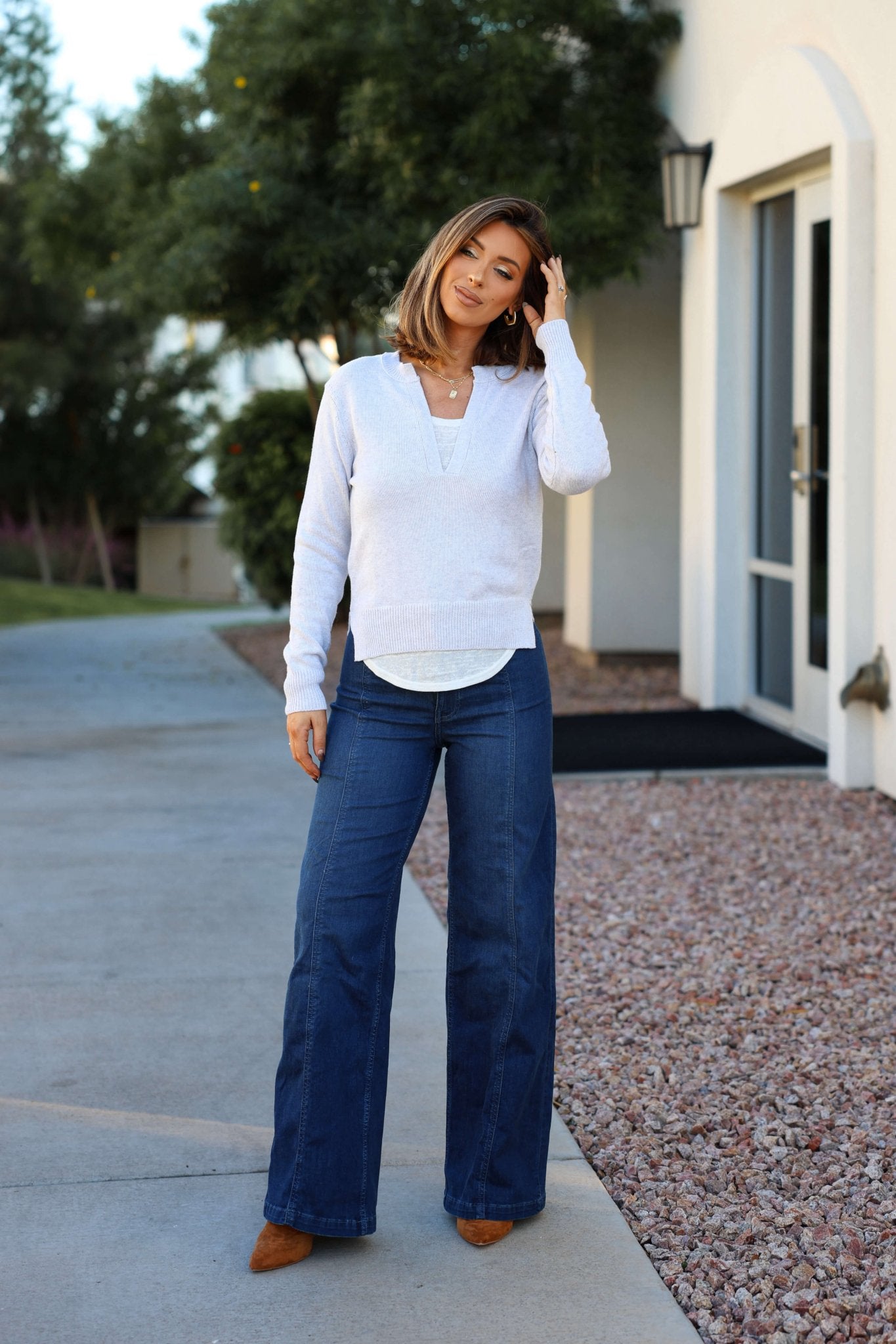A woman stands outdoors in front of a building, wearing a Grey V Neck Ribbed Sweater, wide-leg jeans, and brown shoes.