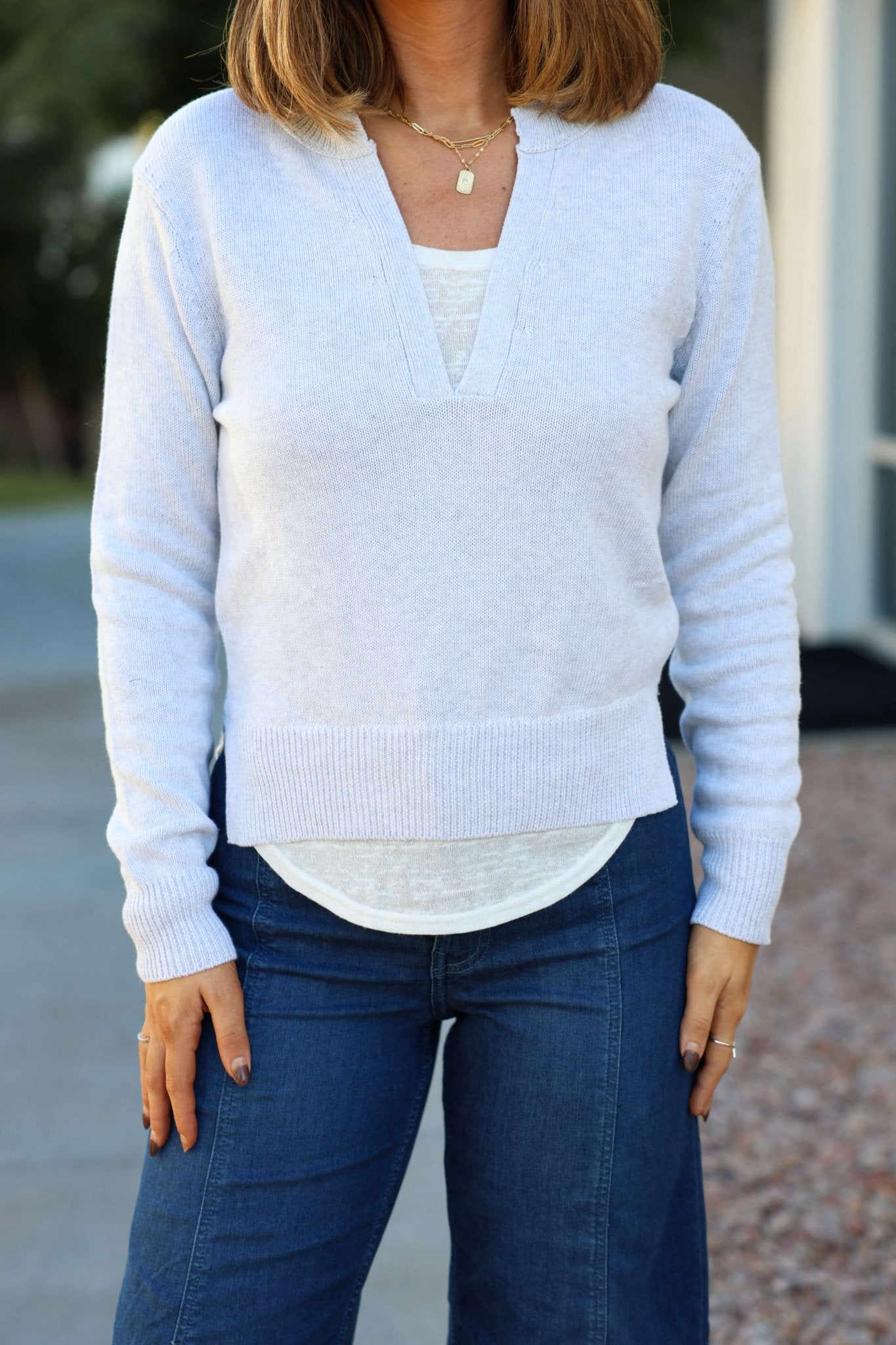 Woman in a Grey V Neck Ribbed Sweater over a white shirt and blue jeans, standing outdoors with relaxed hands.