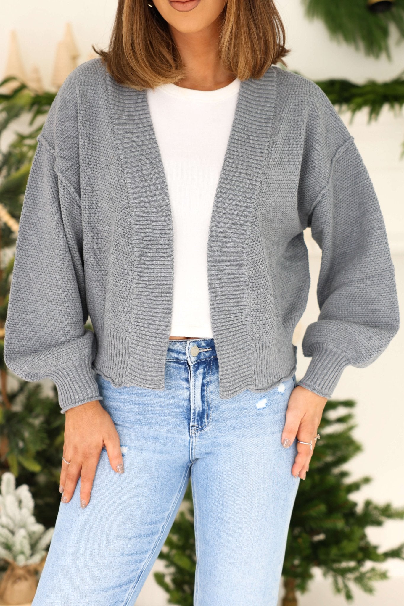 A woman in a Grey Waffle Knit Open Front Cardigan over a white shirt and light blue jeans, standing by a festive green background.