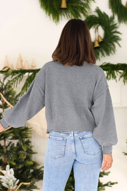 A woman in a Grey Waffle Knit Open Front Cardigan and light blue jeans stands with her back to the camera, among festive greenery.